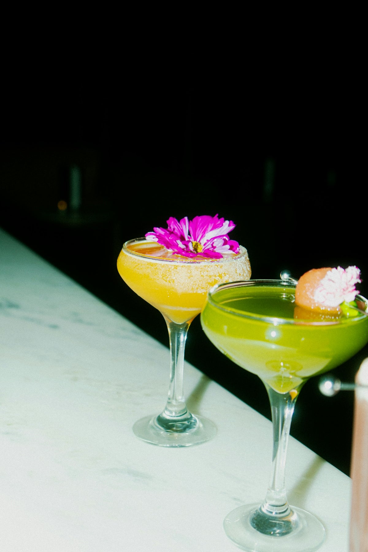 Two colorful cocktails garnished with flowers sit.