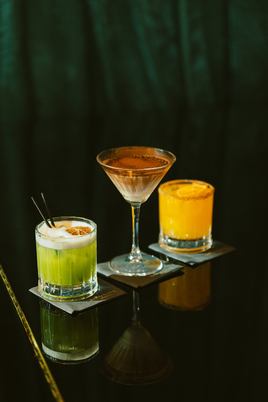 Three colorful cocktails arranged on a dark reflective surface.